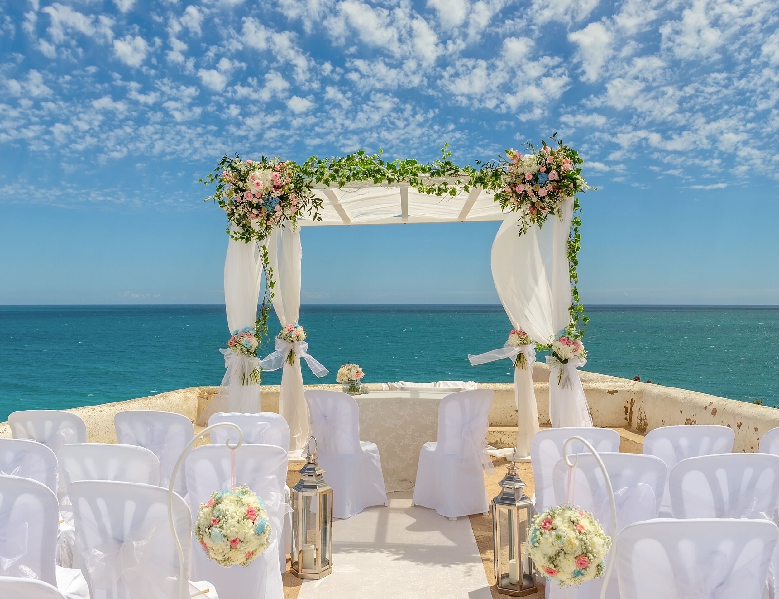 Romantic beach wedding ceremony decor with flowers, drapes, and seating arranged by the sea.